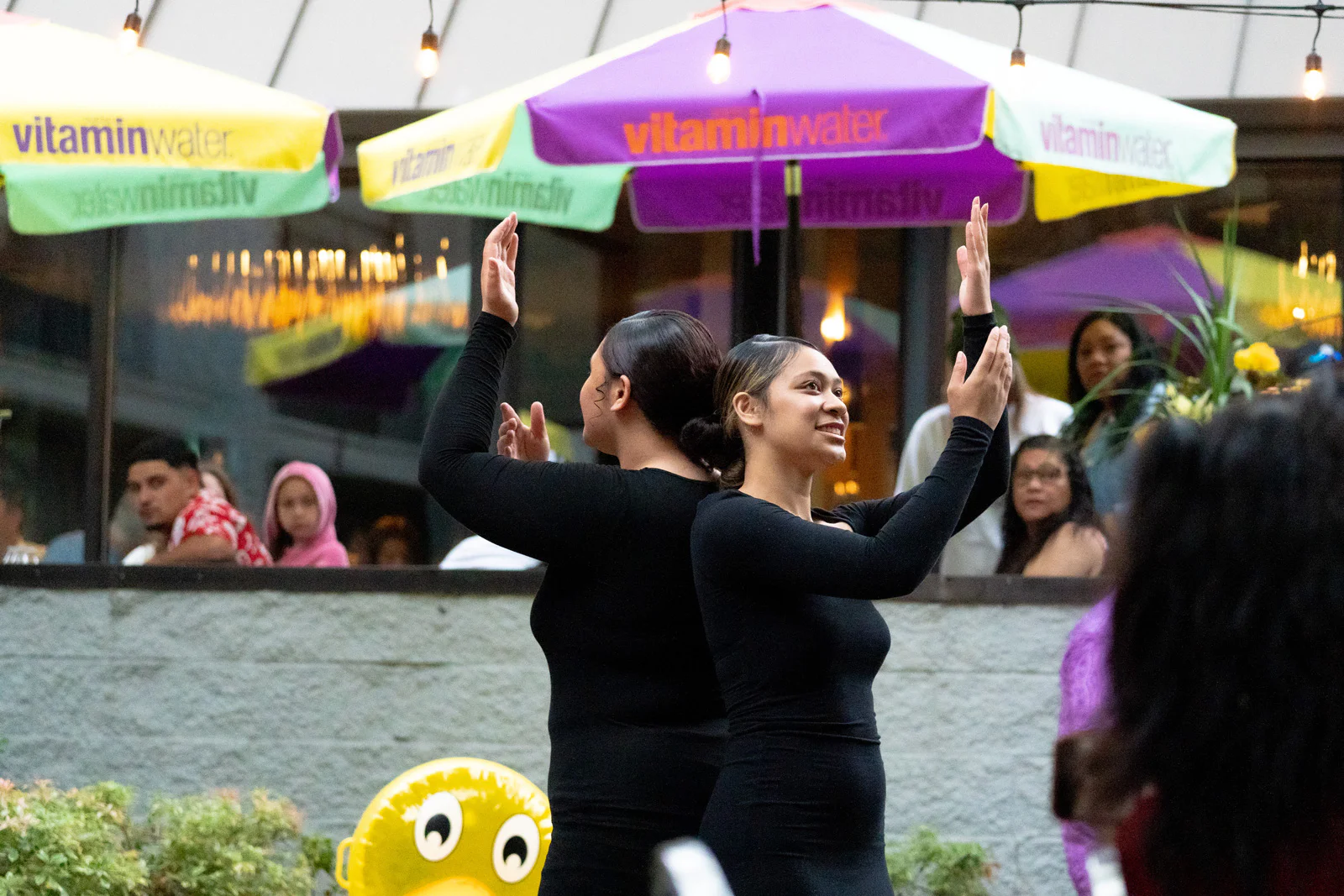 Two people in black outfits perform a synchronized dance outdoors, with colorful umbrellas and string lights above. A small crowd watches in the background.