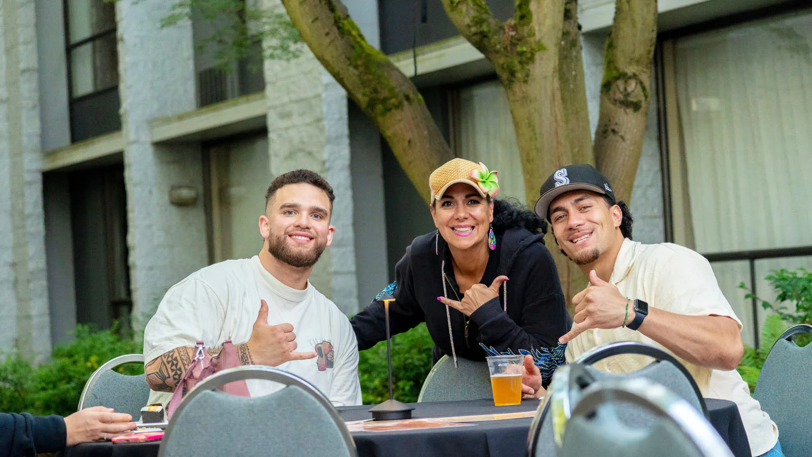 Three people sitting at an outdoor table, smiling and making a shaka hand gesture. They are in a garden setting with trees and a building in the background.