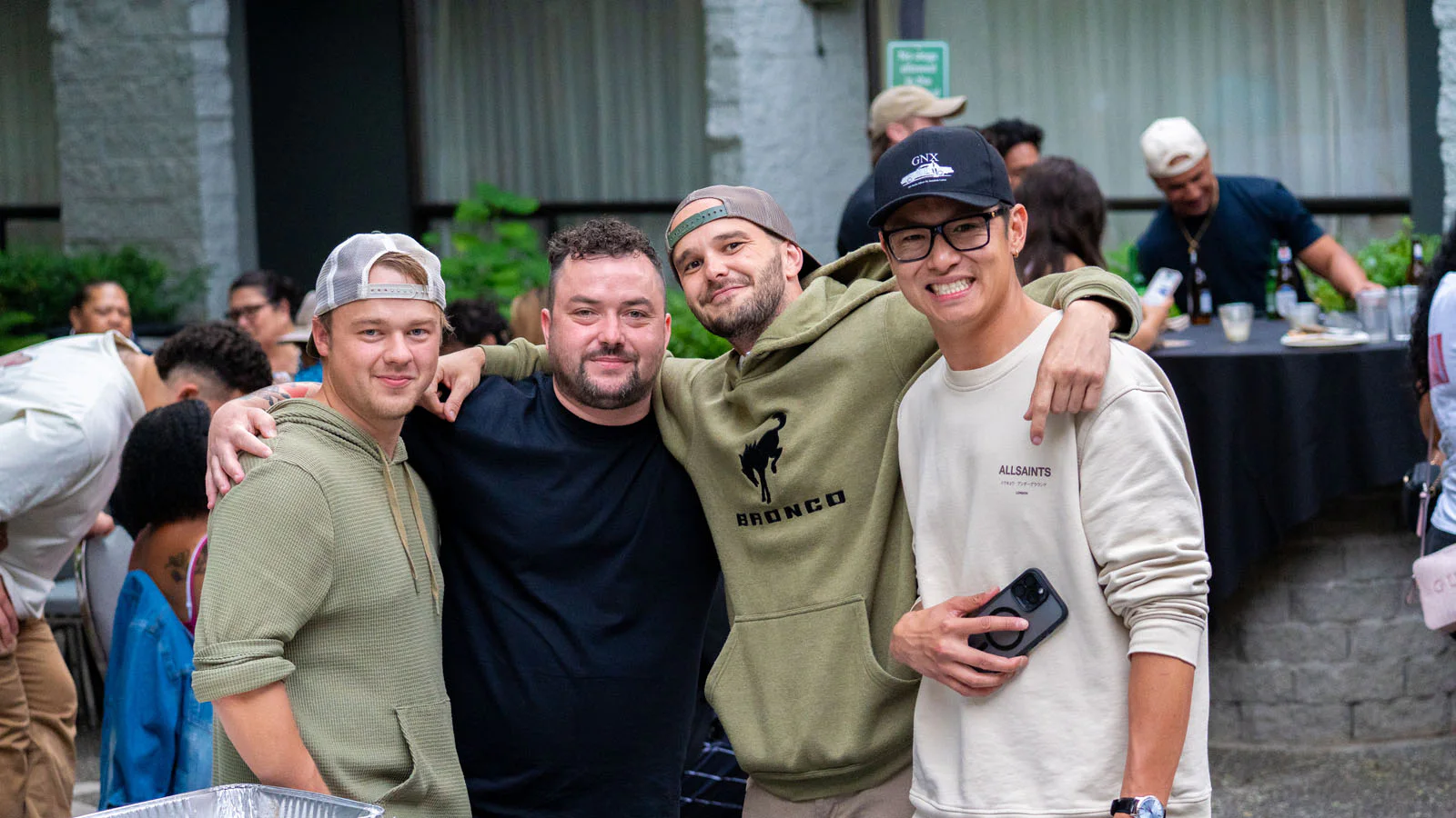 Four men smiling and posing together outdoors at a gathering. They are dressed casually, with others socializing and a table with drinks in the background.