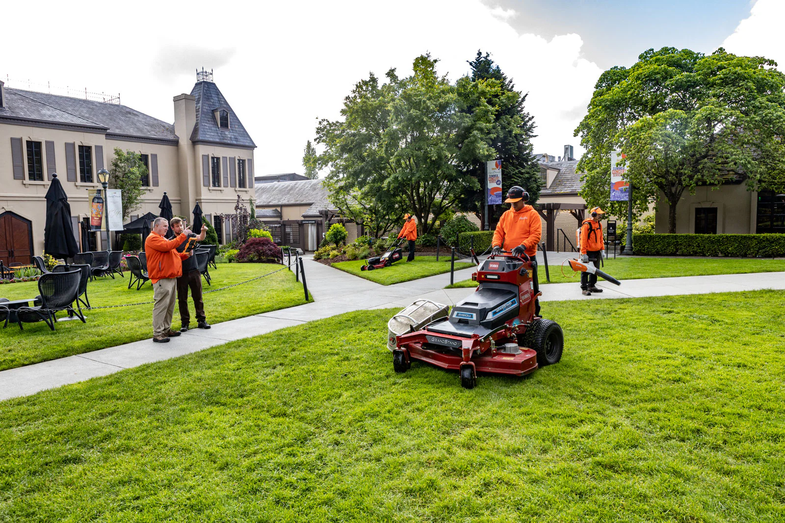 Workers in orange uniforms maintain a landscaped garden outside a large house. One operates a lawnmower, another trims edges, and two discuss plans. Lush, green lawns surround them.