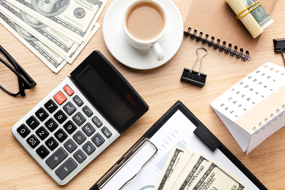 Calculator, glasses, cup of coffee, cash, notepad, binder clip, and calendar organized on a wooden desk. Financial documents and bundled cash suggest planning or budgeting tasks.