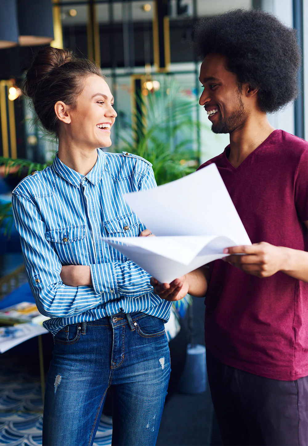 A woman in a striped shirt and jeans smiles while talking to a man in a maroon shirt holding papers. They stand in a modern, well-lit office space with plants.