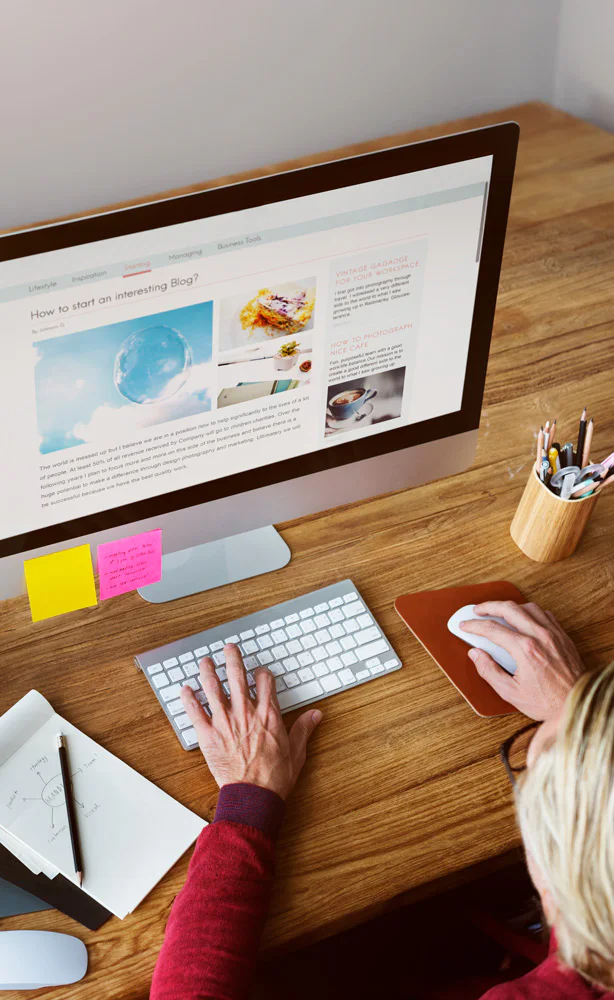 Person using a computer with a blog on the screen. Desk has sticky notes, a notebook with a diagram, and a cup of pens. The blog entry title is 